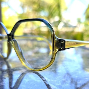 May include: A pair of oversized, olive-green eyeglasses with a gradient effect. The frames have a thick, angular design, and the temples are a lighter shade of green. The glasses are set against a blurred, natural background.