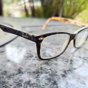 May include: Ray-Ban spectacles with a dark brown and tortoiseshell pattern frame. The glasses have a rectangular shape and clear lenses. The Ray-Ban logo is visible on the temple.