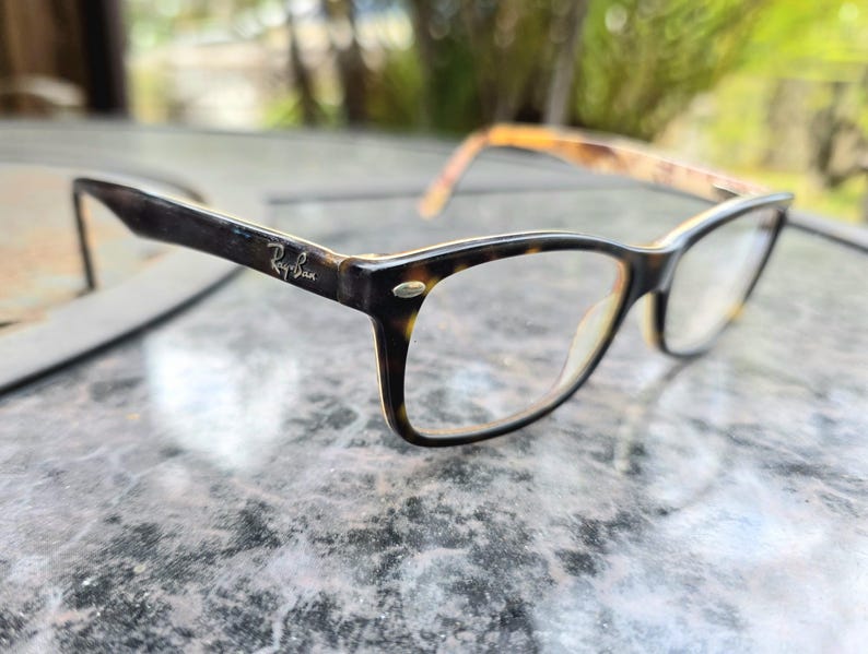 May include: Ray-Ban spectacles with a dark brown and tortoiseshell pattern frame. The glasses have a rectangular shape and clear lenses. The Ray-Ban logo is visible on the temple.