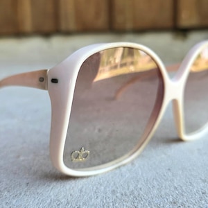 May include: Oversized, light pink sunglasses with gradient lenses. The frames have a rounded shape and a gold crown emblem on the lower left lens. The arms are a matching light pink color.