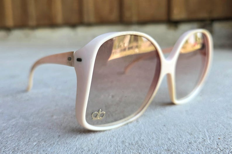 May include: Oversized, light pink sunglasses with gradient lenses. The frames have a rounded shape and a gold crown emblem on the lower left lens. The arms are a matching light pink color.