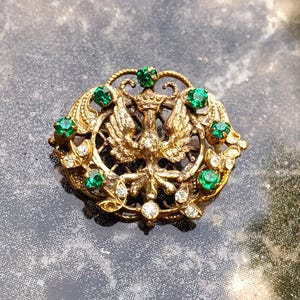 Könnte beinhalten: Vintage goldfarbene Brosche mit einem detaillierten Design eines doppelköpfigen Adlers, verziert mit grünen und klaren Edelsteinen. Die Brosche hat einen verzierten, durchbrochenen Rahmen mit floralen Akzenten, der einen klassischen, eleganten Look erzeugt.