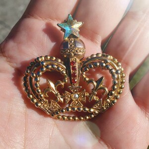May include: A gold crown brooch with a star-shaped jewel on top and red and clear rhinestones. The crown is detailed with intricate designs and beading.
