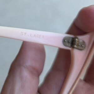 May include: Close-up of a pair of light pink eyeglasses. The text "ST. LAREL" is printed on the arm of the glasses. The hinge is a tarnished brass color. The glasses are held by a hand.