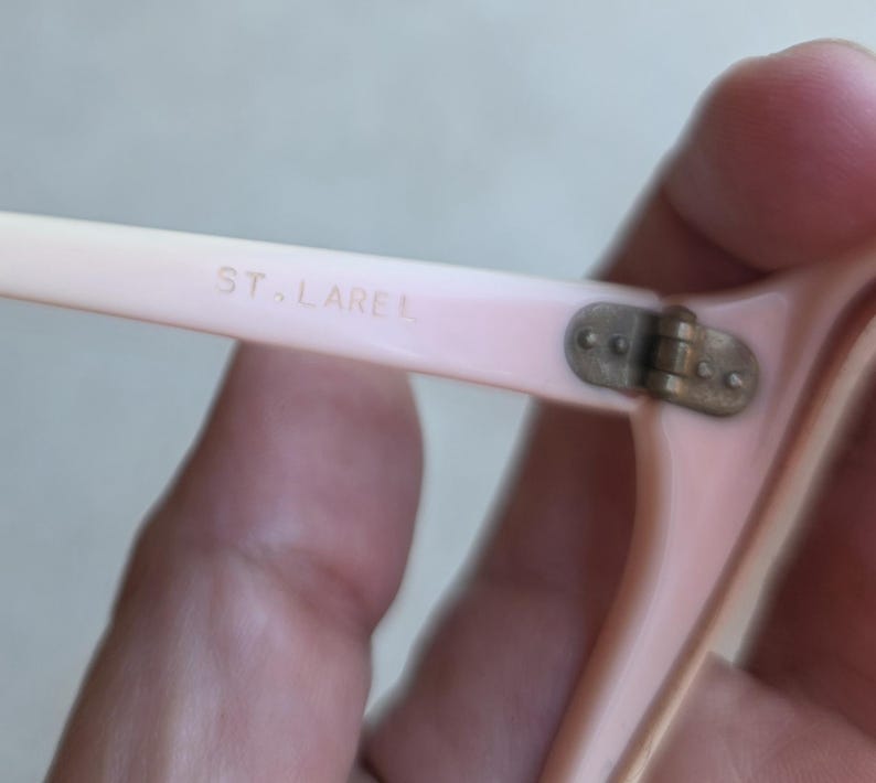 May include: Close-up of a pair of light pink eyeglasses. The text "ST. LAREL" is printed on the arm of the glasses. The hinge is a tarnished brass color. The glasses are held by a hand.