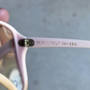 May include: Close-up of light pink eyeglasses with a rectangular frame. The text "MADE IN ITALY 04-2B3" is printed on the temple arm in gold lettering. The glasses have a classic design.