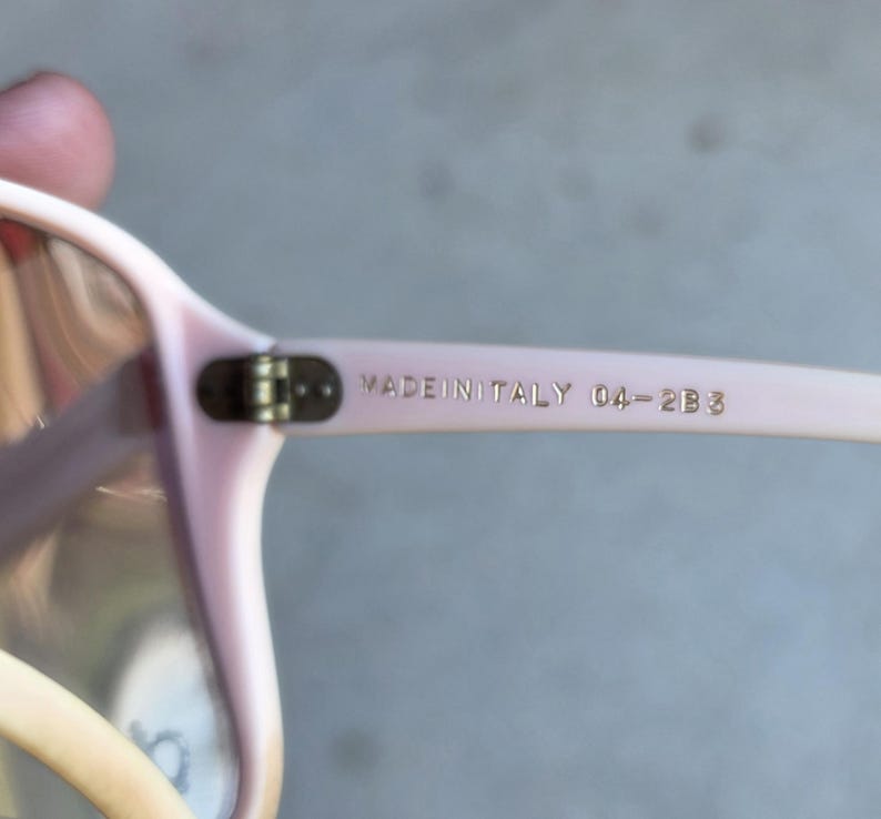May include: Close-up of light pink eyeglasses with a rectangular frame. The text "MADE IN ITALY 04-2B3" is printed on the temple arm in gold lettering. The glasses have a classic design.