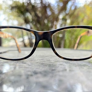 May include: A pair of eyeglasses featuring a tortoiseshell design on the frames. The frames are a mix of dark brown and tan, with clear lenses. The arms of the glasses are light brown. The glasses are resting on a reflective surface.