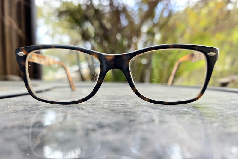 May include: A pair of eyeglasses featuring a tortoiseshell design on the frames. The frames are a mix of dark brown and tan, with clear lenses. The arms of the glasses are light brown. The glasses are resting on a reflective surface.