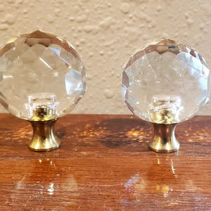 May include: Two clear, faceted, spherical glass knobs with polished gold-tone metal bases. Each knob features a multi-faceted surface that reflects light, attached to a flared, circular metal pedestal base designed for mounting on furniture or doors.