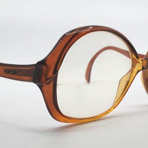 May include: Vintage oversized eyeglasses with a brown tortoiseshell frame. The glasses have a large, rounded shape with clear lenses and curved temples. The frame has a subtle decorative detail near the hinges.