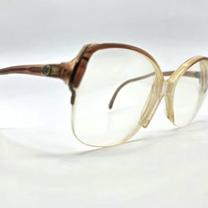 May include: Vintage eyeglasses with large, clear lenses and a brown and tan frame. The frames have a unique, angular shape and a decorative emblem on the temples. The glasses are set against a white background.