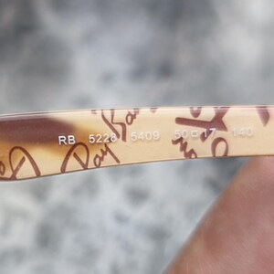 May include: Close-up of the arm of a pair of Ray-Ban sunglasses. The arm is tan with brown script and the numbers RB 5228 5409 50-17-140. Sunglasses are a popular fashion accessory.