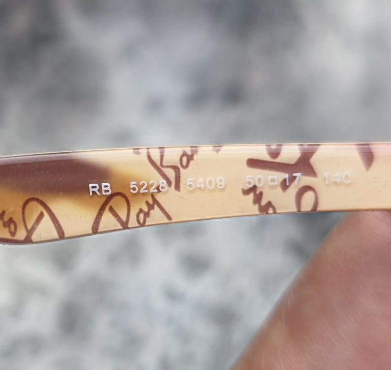 May include: Close-up of the arm of a pair of Ray-Ban sunglasses. The arm is tan with brown script and the numbers RB 5228 5409 50-17-140. Sunglasses are a popular fashion accessory.