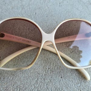 May include: Vintage oversized sunglasses with a cream-colored frame and brown lenses. The glasses have a unique shape and feature the text "MADE IN ITALY" on the arm. A small gold-colored emblem is visible on the left lens.