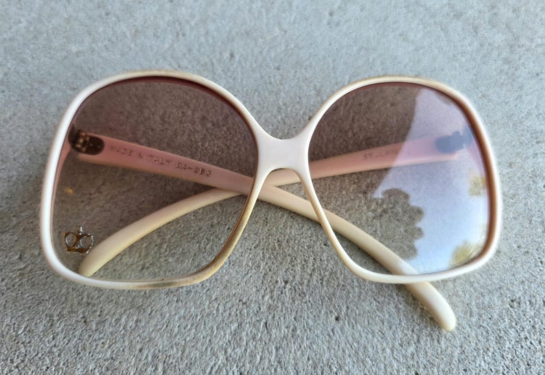 May include: Vintage oversized sunglasses with a cream-colored frame and brown lenses. The glasses have a unique shape and feature the text "MADE IN ITALY" on the arm. A small gold-colored emblem is visible on the left lens.