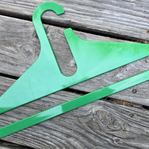 Puede incluir: Una percha de plástico verde brillante. La percha tiene un gancho curvado y un cuerpo ancho y plano con una abertura rectangular. La percha está hecha de un material plástico liso y sólido.