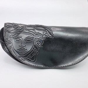 Op de afbeelding: Zwarte leren heuptas met een Medusa-hoofdontwerp aan de voorkant. De tas heeft een gebogen vorm en een zwarte riem. De Medusa-kop is in het leer ge&euml;mbosseerd.