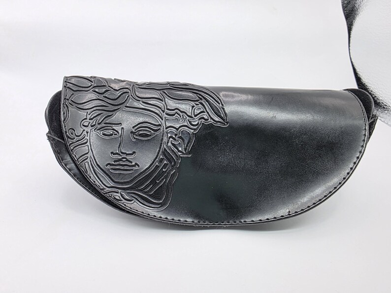 Op de afbeelding: Zwarte leren heuptas met een Medusa-hoofdontwerp aan de voorkant. De tas heeft een gebogen vorm en een zwarte riem. De Medusa-kop is in het leer ge&euml;mbosseerd.