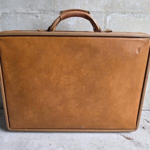 May include: A rectangular tan leather suitcase with a structured frame and a top handle. The exterior features a smooth, solid-colored surface with a small embossed logo detail on the upper right corner and a reinforced border trim.