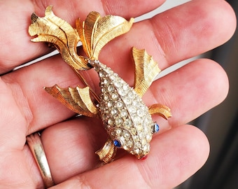 Puede incluir: Broche de pez dorado con un cuerpo incrustado con pedrería transparente. El pez tiene detalles azules y rojos para los ojos y la boca. El broche presenta aletas y cola texturizadas, y un cierre de alfiler en la parte posterior.