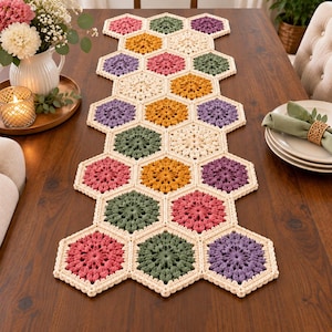 Puede incluir: Un camino de mesa de ganchillo compuesto por motivos hexagonales interconectados en varios colores, incluyendo crema, amarillo mostaza, verde salvia, rosa viejo y lavanda. El camino presenta un patrón geométrico repetitivo con un borde crema.