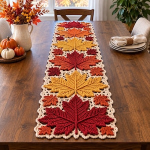 May include: A rectangular crochet table runner featuring a repeating pattern of large and small maple leaves in shades of red, orange, and yellow, bordered by a cream-coloured scalloped edge, laid flat on a dark brown wooden dining table surface.