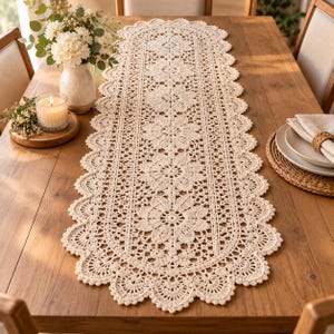 Peut inclure: Un chemin de table en crochet de couleur crème, de forme allongée, présentant un motif floral répétitif au centre, bordé d'une dentelle ajourée complexe et d'un bord festonné. Le textile est réalisé avec un fil fin au design symétrique.