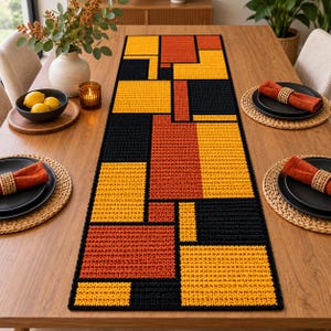 Puede incluir: Un camino de mesa rectangular con un patrón geométrico de bloques en negro, amarillo mostaza y naranja quemado. El camino tiene una apariencia tejida y texturizada con líneas limpias y bordes definidos, creando una cuadrícula estructurada.