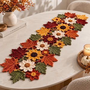 Pode incluir: Um caminho de mesa decorativo e comprido feito de elementos de croché, com um padrão repetitivo de folhas de ácer em cores outonais em tons de vermelho, laranja e verde, intercaladas com flores brancas e amarelas.