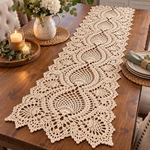 May include: A long, rectangular cream-colored crochet table runner featuring an intricate repeating pineapple-inspired lace pattern with scalloped edges, crafted from fine thread and laid flat across the center of a wooden dining table.