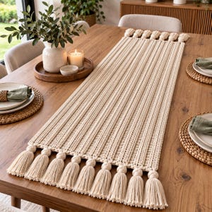 Puede incluir: Un camino de mesa de macramé de color crema, largo y rectangular, con un patrón de cordón texturizado y acabado con una hilera de borlas anudadas decorativas en ambos extremos. El camino está hecho de cordón de algodón grueso y plano.