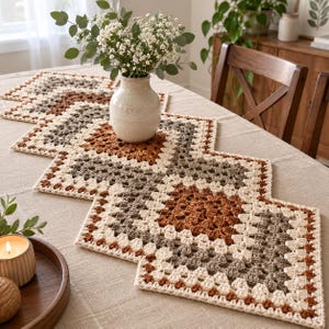 Könnte beinhalten: Ein gehäkelter Tischläufer mit einer Reihe verbundener quadratischer Motive in einer Farbpalette aus Creme, Hellbraun und Rostorange. Der Läufer ist in einem Zickzackmuster auf einem Tisch ausgelegt, mit einer Keramikvase mit weißen Blumen.