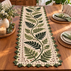 May include: A rectangular crochet table runner featuring an off-white base with a central winding vine pattern adorned with various shades of green fern leaves. The edges are finished with a decorative scalloped border in dark and light green.