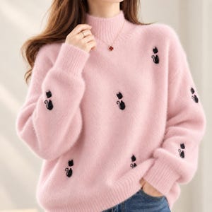 May include: A soft, fuzzy, light pink mock neck jumper featuring long sleeves and ribbed cuffs. The jumper is decorated with small, embroidered black cat silhouettes scattered across the front and sleeves. The fabric has a textured, plush appearance.