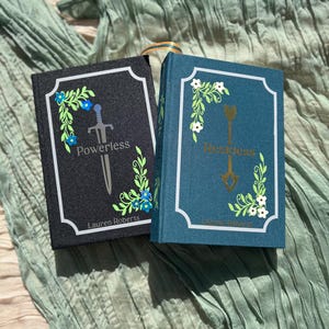 May include: Two hardcover books, one black and one teal, with white borders and floral designs. The black book reads "Powerless" with a sword illustration, and the teal book reads "Reckless" with an arrow illustration. Both books have the author's name.