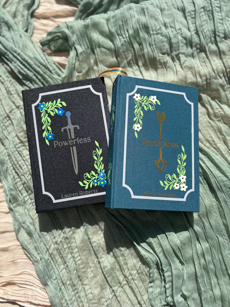 May include: Two hardcover books, one black and one teal, with white borders and floral designs. The black book reads "Powerless" with a sword illustration, and the teal book reads "Reckless" with an arrow illustration. Both books have the author's name.