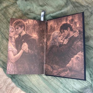 May include: Open book with sepia-toned illustrations. One page shows a couple dancing, the other depicts a dramatic scene with a woman and man embracing, and a fallen figure. A silver bookmark is visible.