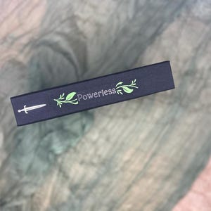 May include: A dark blue box with the word "Powerless" in green script, accompanied by a sword and leaf design. The box is rectangular and appears to be made of cardboard or a similar material. The background is a textured, light-colored fabric.