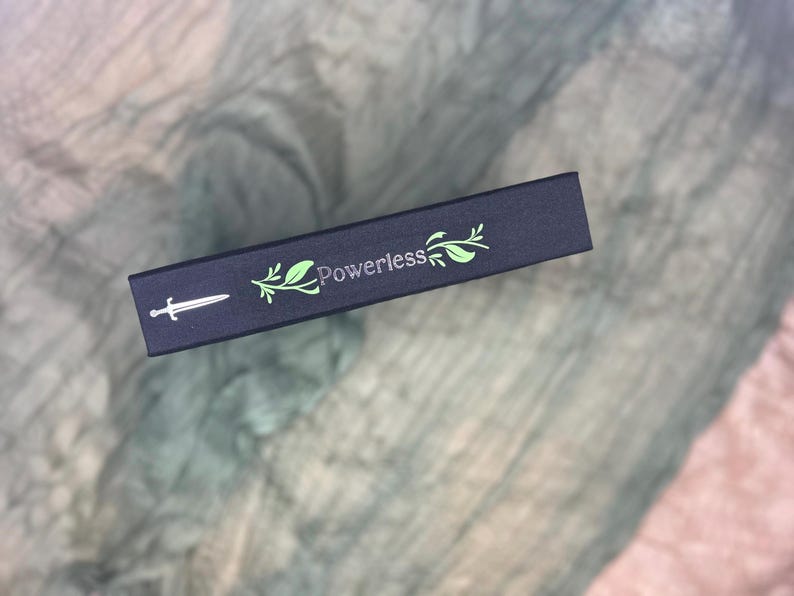 May include: A dark blue box with the word "Powerless" in green script, accompanied by a sword and leaf design. The box is rectangular and appears to be made of cardboard or a similar material. The background is a textured, light-colored fabric.