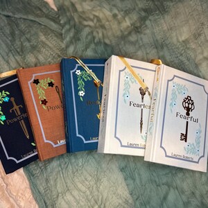 May include: A collection of hardcover books with decorative floral and key designs. The books have titles like "Powerless" and "Fearful" with gold ribbon bookmarks. The covers are in shades of navy, brown, and white, with the author's name at the bottom.