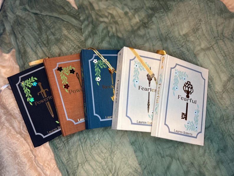 May include: A collection of hardcover books with decorative floral and key designs. The books have titles like "Powerless" and "Fearful" with gold ribbon bookmarks. The covers are in shades of navy, brown, and white, with the author's name at the bottom.