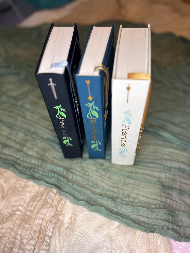 May include: Three hardcover books with decorative covers. The books are in navy blue, blue, and white. Each book has gold lettering and a decorative design. The spines display the titles "Powerless", "Reckless", and "Fearless". Each book has a ribbon bookmark.