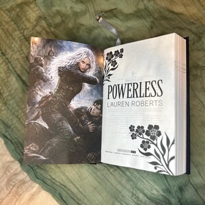 May include: An open book with the title "POWERLESS" by Lauren Roberts. The cover art features a woman with long white hair in a dark outfit, and a man. The right page has floral illustrations.