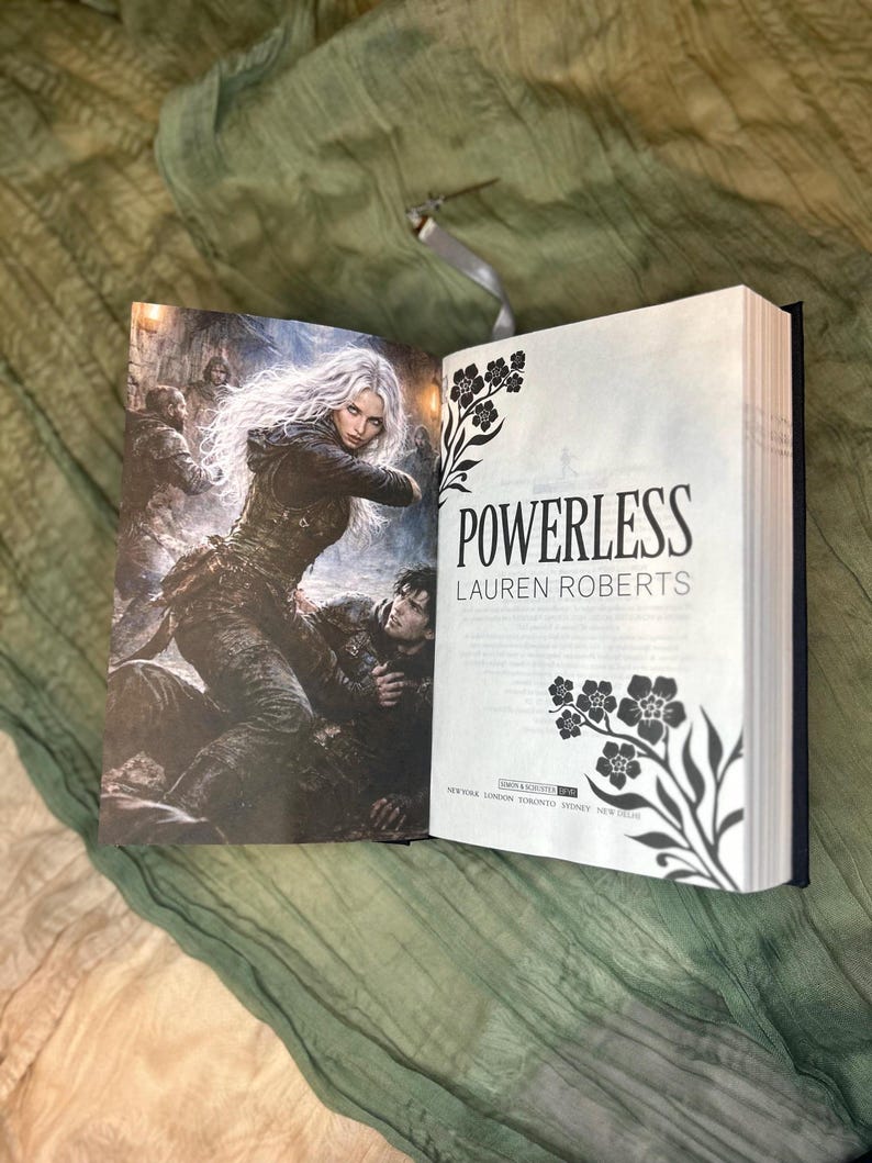 May include: An open book with the title "POWERLESS" by Lauren Roberts. The cover art features a woman with long white hair in a dark outfit, and a man. The right page has floral illustrations.
