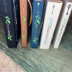 May include: A collection of hardcover books with decorative covers. The books are in various colors, including black, brown, and blue, with gold foil lettering and leaf designs. Each book has a matching bookmark with a charm. The titles include "Powerless", "Powerful", "Reckless", and "Fearless".