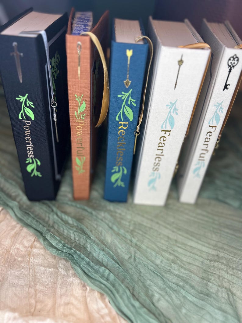 May include: A collection of hardcover books with decorative covers. The books are in various colors, including black, brown, and blue, with gold foil lettering and leaf designs. Each book has a matching bookmark with a charm. The titles include "Powerless", "Powerful", "Reckless", and "Fearless".