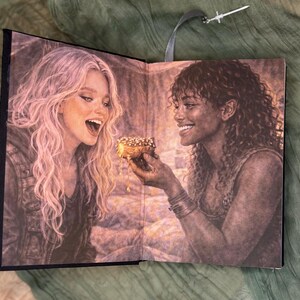 May include: Open book with a detailed illustration of two women sharing a pastry. One woman with long blonde hair is laughing, while the other, with dark curly hair, offers the pastry. A silver sword-shaped bookmark hangs from the top.