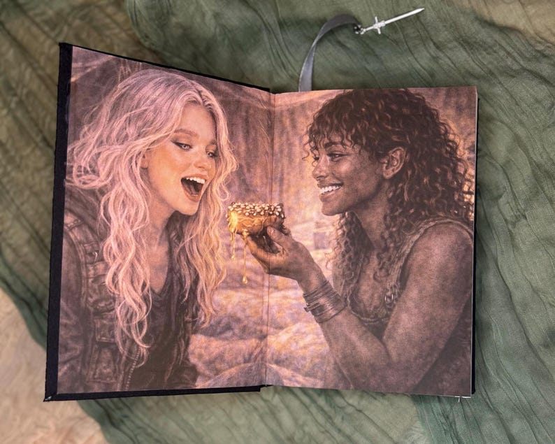 May include: Open book with a detailed illustration of two women sharing a pastry. One woman with long blonde hair is laughing, while the other, with dark curly hair, offers the pastry. A silver sword-shaped bookmark hangs from the top.