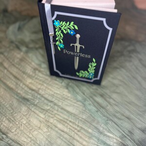 May include: A hardcover book with a black cover featuring the word "Powerless" and a sword illustration. The cover is decorated with floral and leaf designs in blue and green. A silver ribbon bookmark is attached.